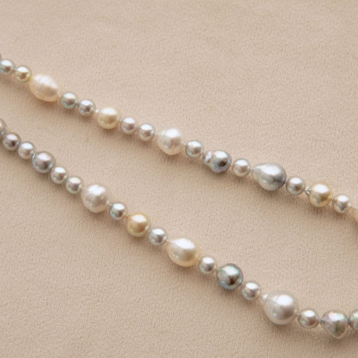 A strand of mixed Australian South Sea and Akoya pearls in various sizes and colors, including cream, white, and iridescent grey, laid out diagonally on a textured beige background.