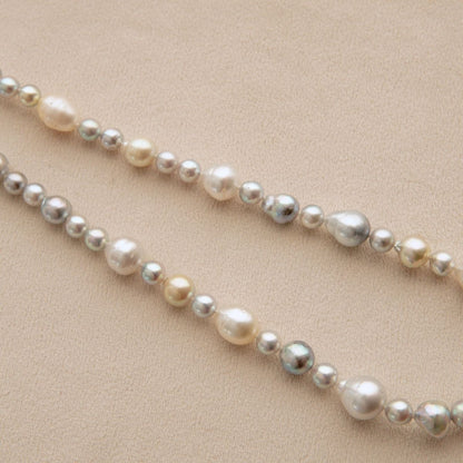 A strand of mixed Australian South Sea and Akoya pearls in various sizes and colors, including cream, white, and iridescent grey, laid out diagonally on a textured beige background.