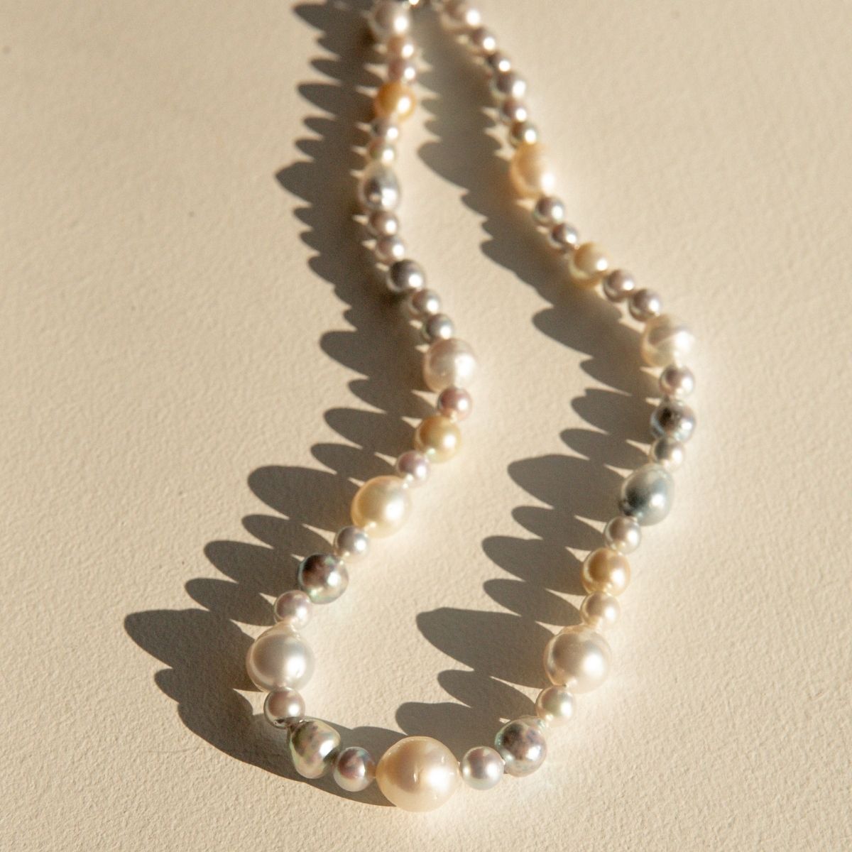 A strand of mixed Australian Saltwater pearls in cream, ivory, and silver tones, arranged in a loose loop on a warm beige surface with sharp sunlight shadows.