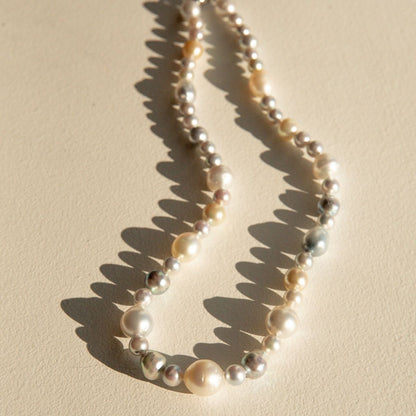 A strand of mixed Australian Saltwater pearls in cream, ivory, and silver tones, arranged in a loose loop on a warm beige surface with sharp sunlight shadows.
