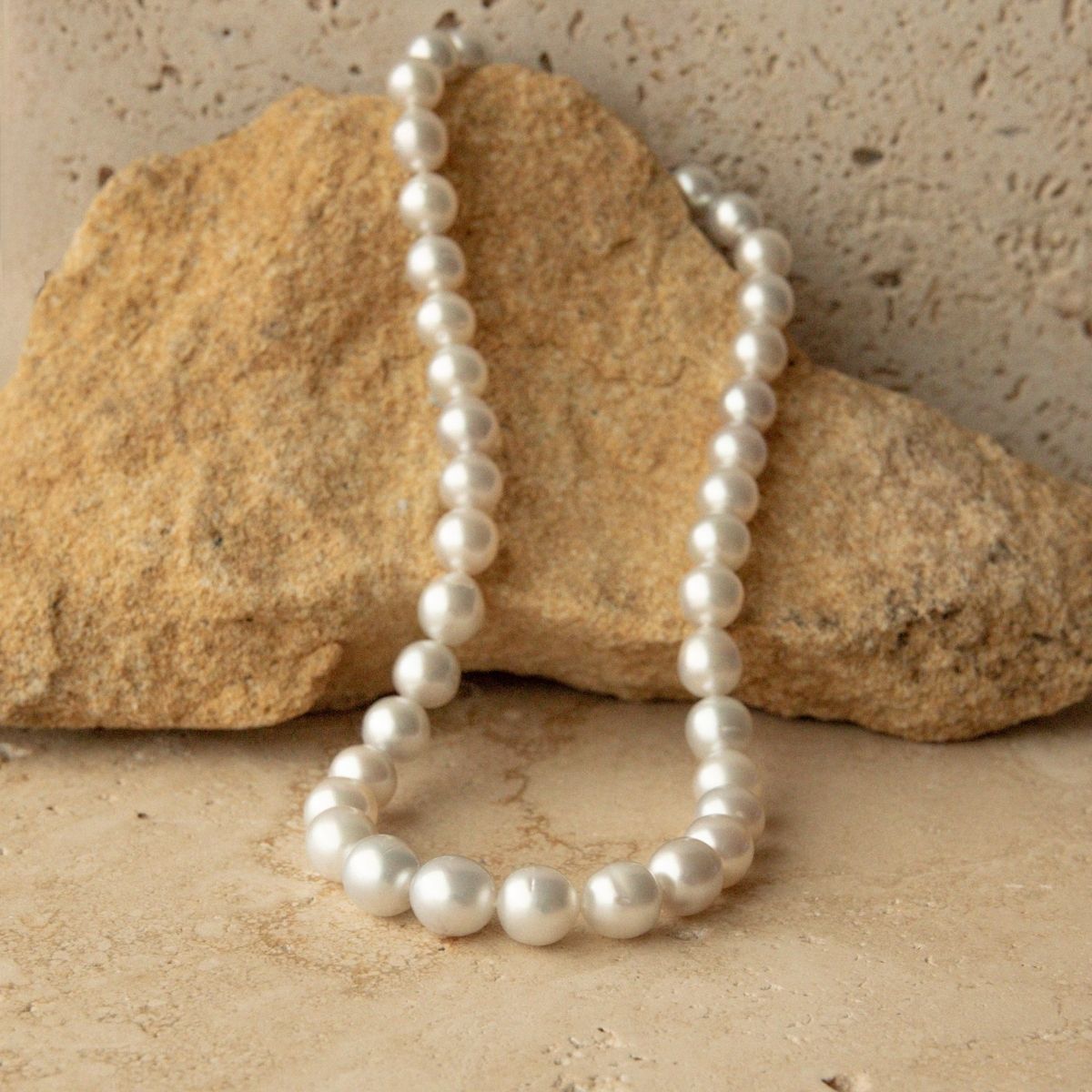 A strand of Australian South Sea pearls draped over a tan rock on a textured beige stone surface.