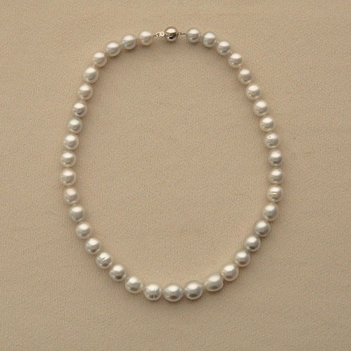 A classic strand of white Australian South Sea pearls arranged in an oval shape on a textured beige background, featuring a silver-toned clasp at the top.