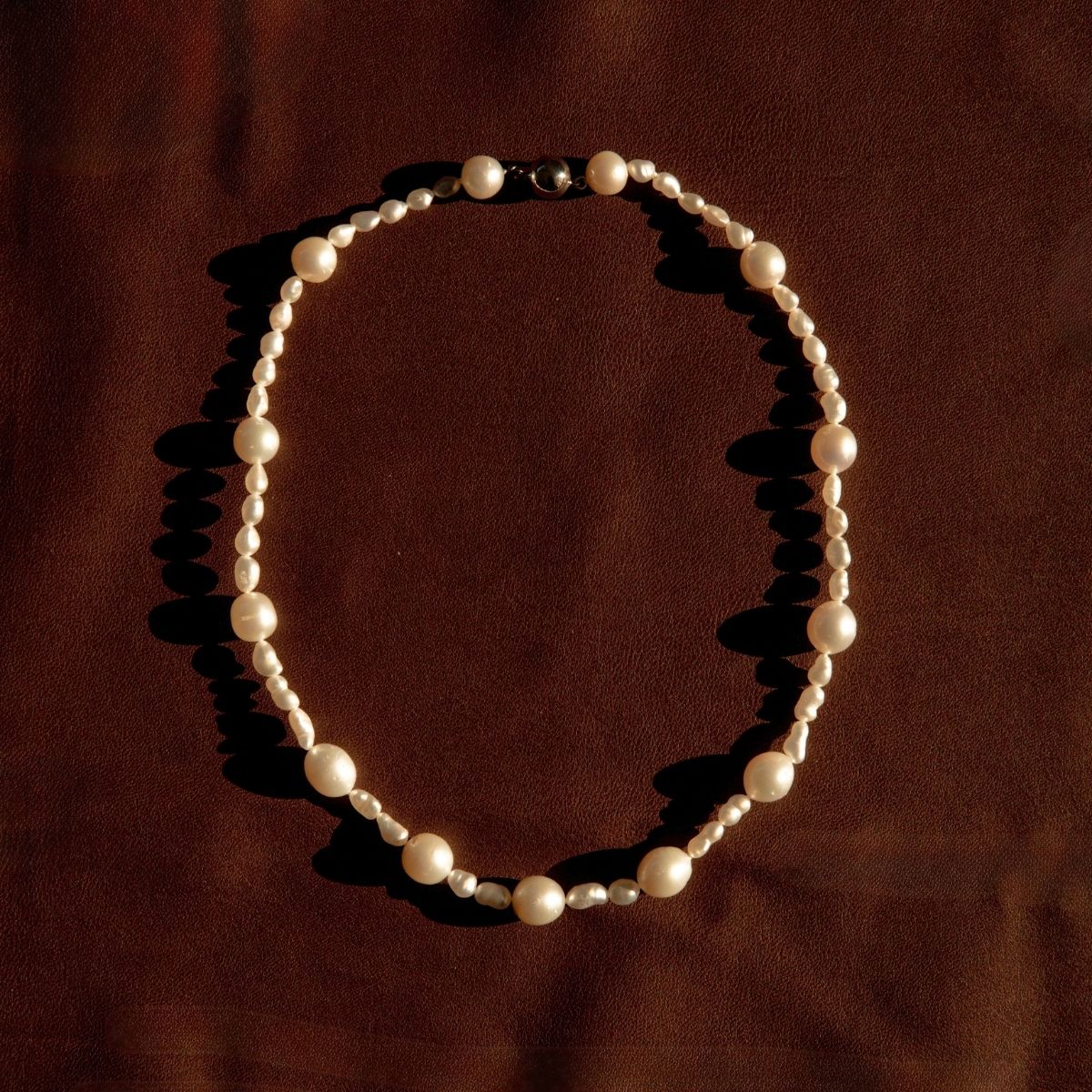 A strand necklace made of Keshi and South Sea pearls, arranged in an oval shape on a dark brown textured fabric background.