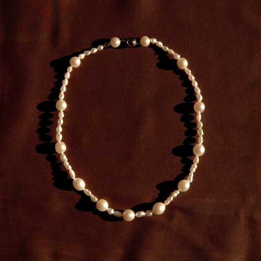 A strand necklace made of Keshi and South Sea pearls, arranged in an oval shape on a dark brown textured fabric background.