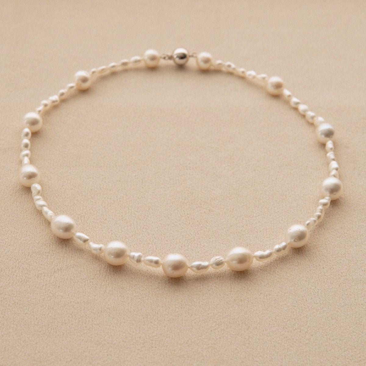 A delicate pearl strand necklace with alternating large and small white Keshi and South Sea pearls on a warm beige textured background.