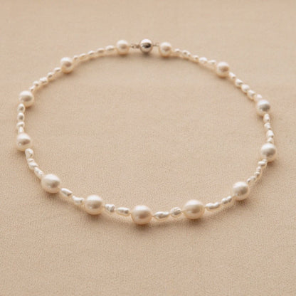 A delicate pearl strand necklace with alternating large and small white Keshi and South Sea pearls on a warm beige textured background.