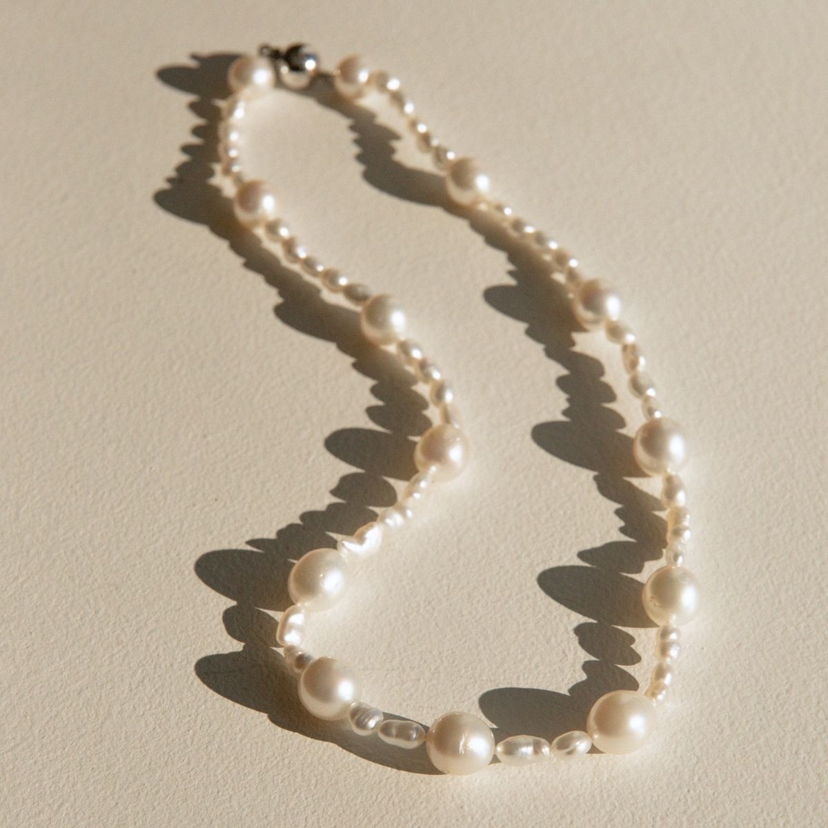 An Australian South Sea & Keshi pearl necklace with mixed pearl sizes, styled on a textured beige surface with dramatic, sharp sunlight casting shadows.