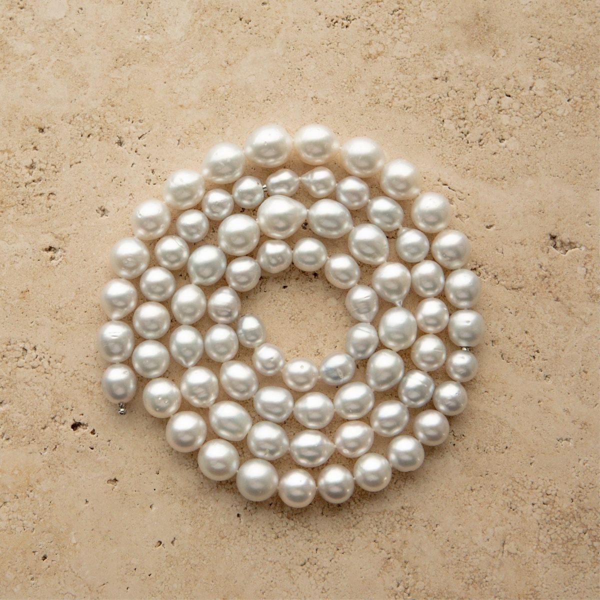 A coiled strand of Australian South Sea pearl beads in drop and semi drop shapes, arranged in a circle on a textured beige stone surface.