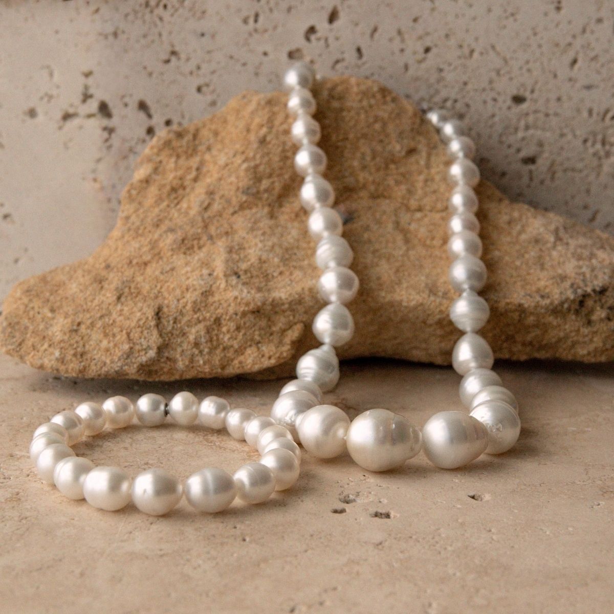 A strand of white South Sea pearls draped over a natural tan rock, with a matching pearl bracelet placed in front on a textured beige stone surface.