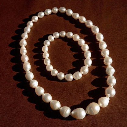 A set of white baroque pearl jewellery pieces, including a long strand necklace and a matching bracelet that can be worn together or separately, arranged in concentric circles on a dark brown textured background.