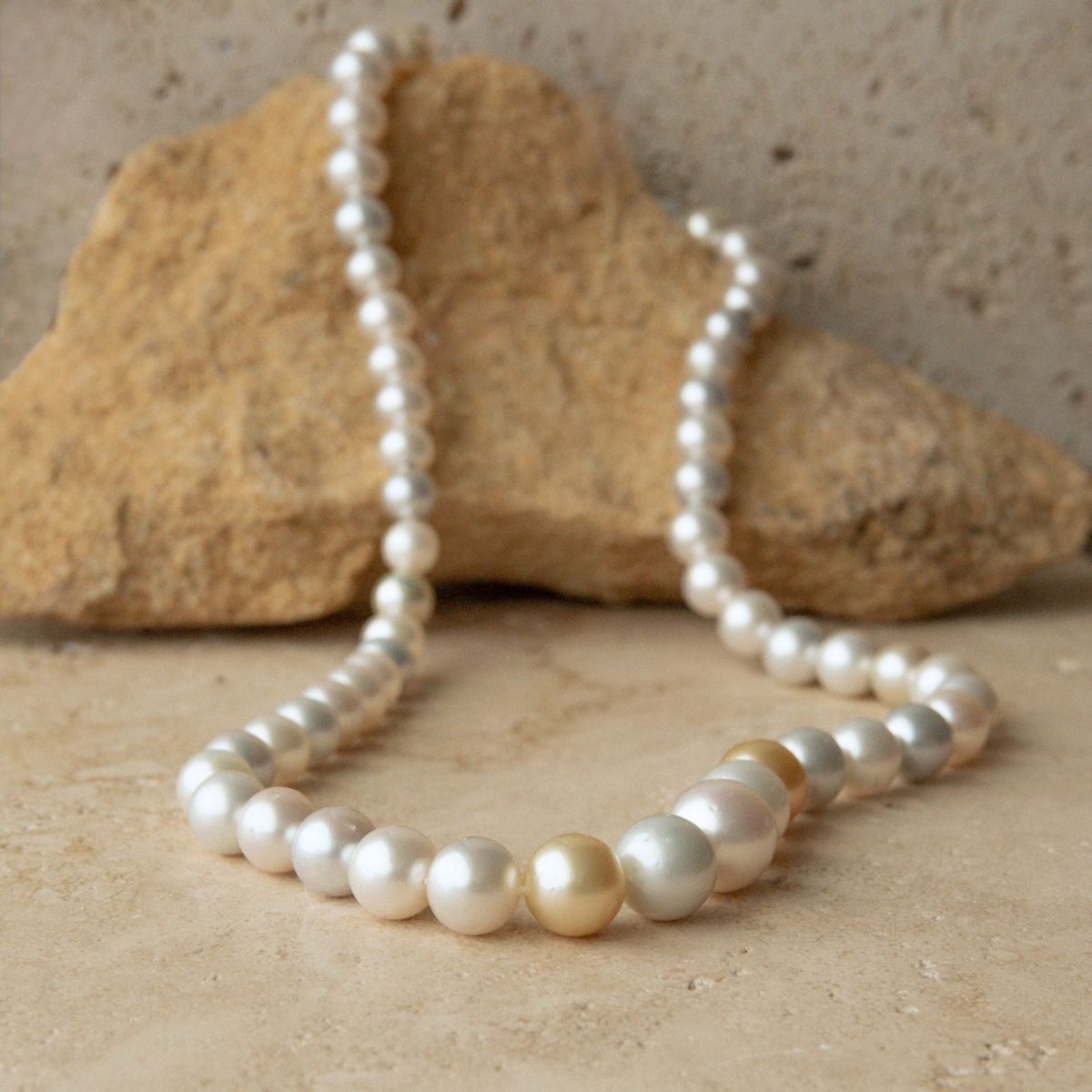 A strand of multicolour Australian South Sea pearls draped in an arc across a beige stone surface, with a larger tan rock behind them.