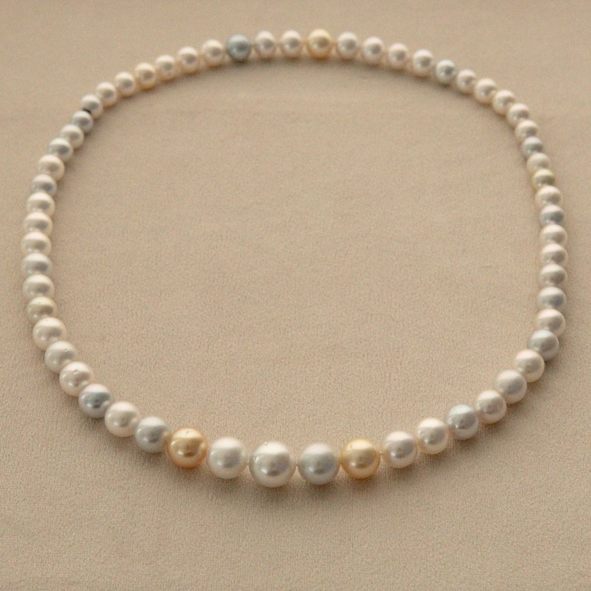 A strand necklace made of multicolour Australian South Sea pearls with a few larger champagne-gold and pale blue/sivler pearls, arranged in a circle on a beige background.
