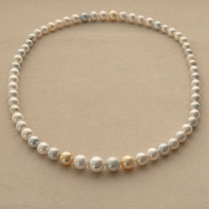 A strand necklace made of multicolour Australian South Sea pearls with a few larger champagne-gold and pale blue/sivler pearls, arranged in a circle on a beige background.