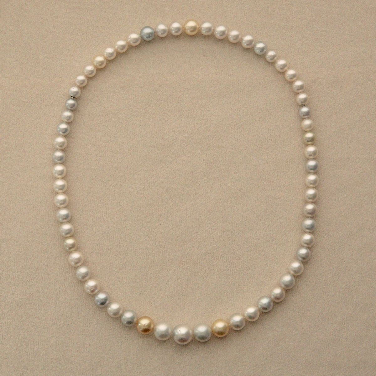 An Australian South Sea pearl necklace with mostly white and silver-gray pearls accented by a few gold and cream pearls, displayed on a neutral beige background.
