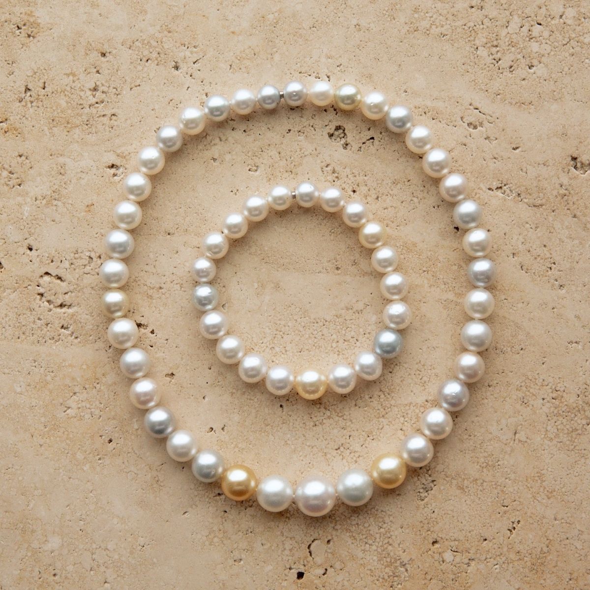 A set of two pearl strand pieces, a longer strand necklace and a matching bracelet, arranged in concentric circles on a textured beige stone surface. The Australian South Sea pearls are predominantly white with a few natural champagne-gold and light gray variations.