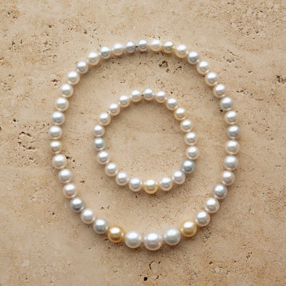 A set of two pearl strand pieces, a longer strand necklace and a matching bracelet, arranged in concentric circles on a textured beige stone surface. The Australian South Sea pearls are predominantly white with a few natural champagne-gold and light gray variations.