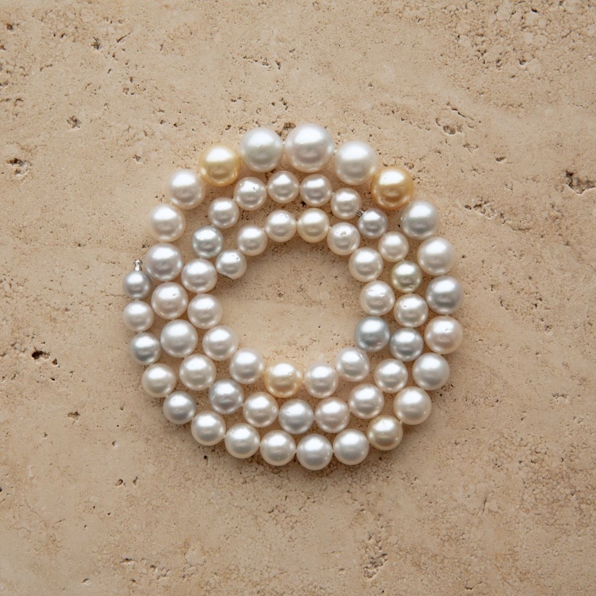 A strand of multicolour Australian South Sea pearls arranged in a loose circle on a textured beige stone surface.