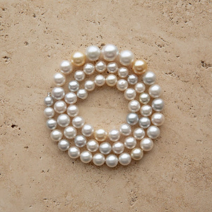 A strand of multicolour Australian South Sea pearls arranged in a loose circle on a textured beige stone surface.