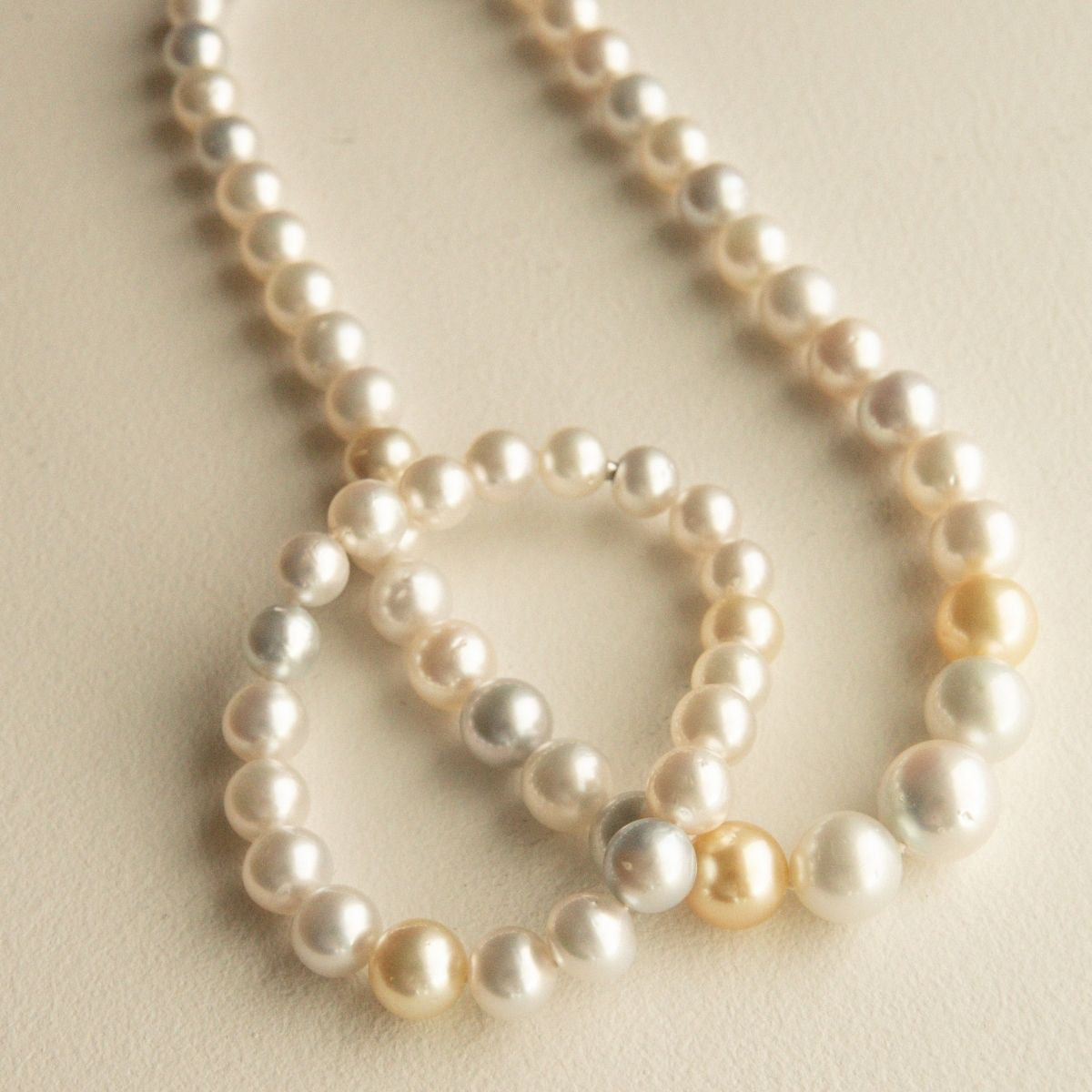 A strand of multicolour Australian South Sea pearls with a few warm cream and pale gold pearls, arranged in a loose circle on a light beige textured background.