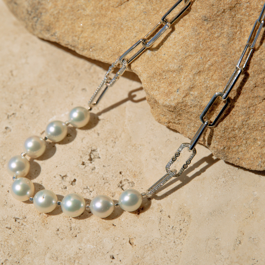 Pearl necklace and white gold paperclip chain on a stone surface.