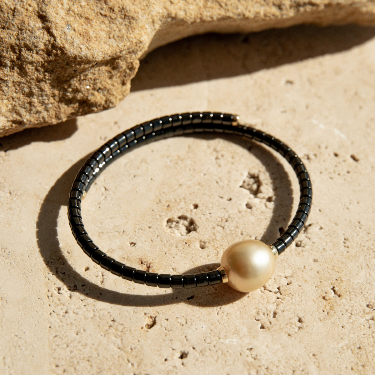 Bracelet with black ceramic links and a gold pearl on a sandy surface.