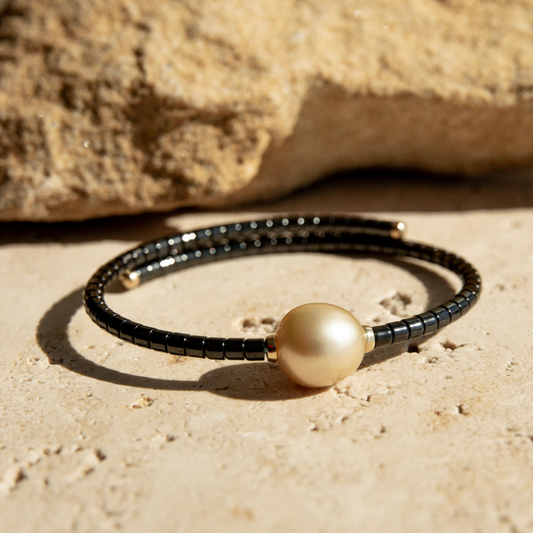Bracelet with black ceramic links and a gold pearl on a sandy surface.