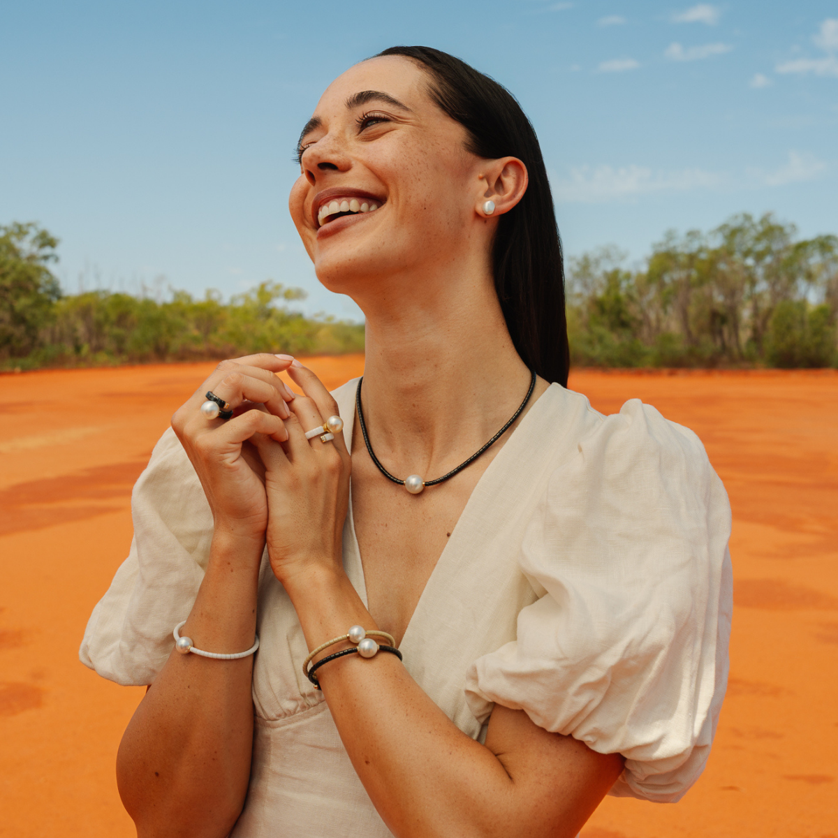 Woman in a white blouse standing on red dirt with trees and blue sky in the background