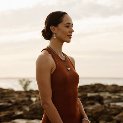 Woman in a rust-colored dress standing on a rocky shore with a cloudy sky wearing pearl hooks earrings.