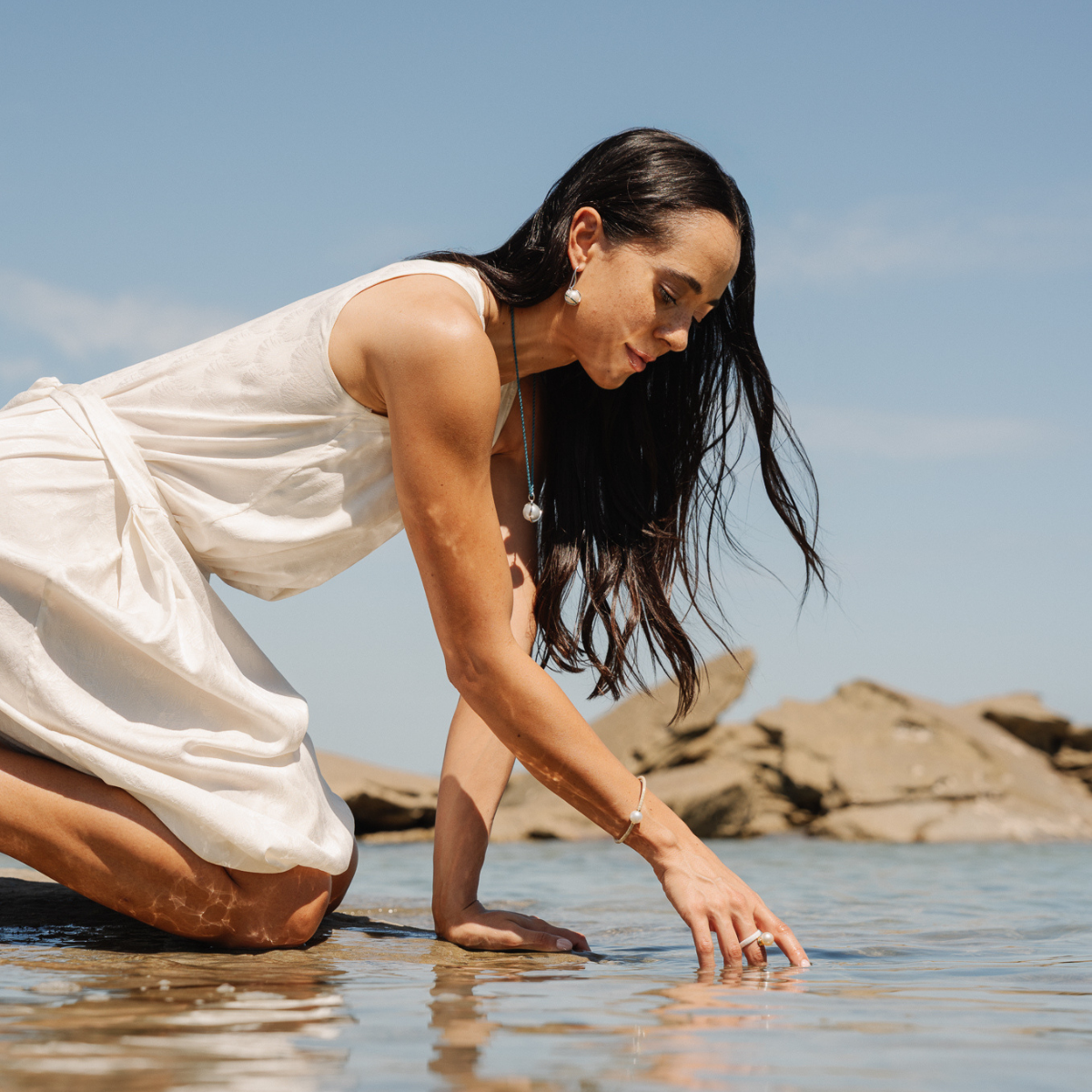 Woman in a white dress touching water on a beach with rocks and clear sky wearing an Australian South Sea pearl pendant.