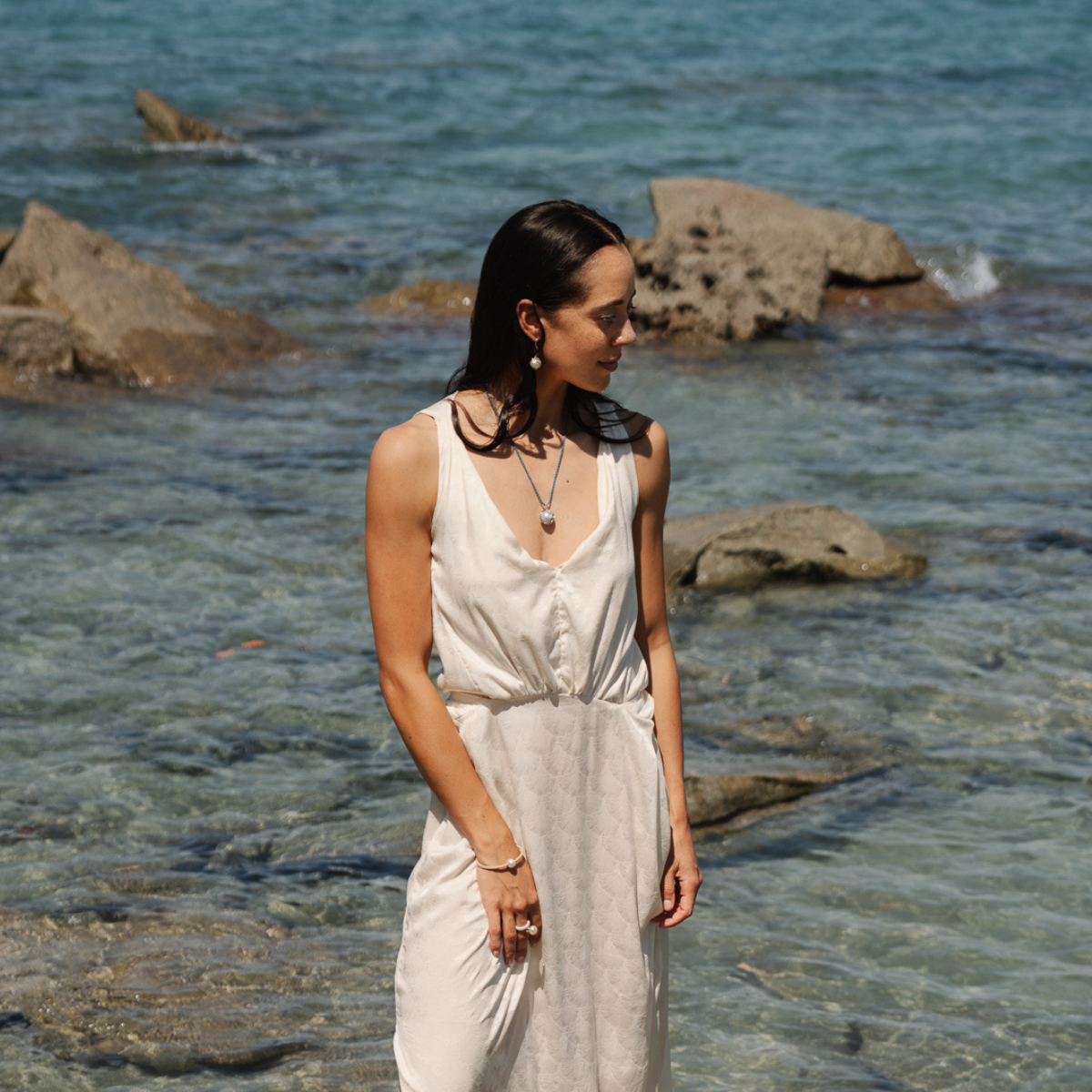 Woman in a white dress standing by the water with rocks in the background wearing a Australian South Sea pearl pendant