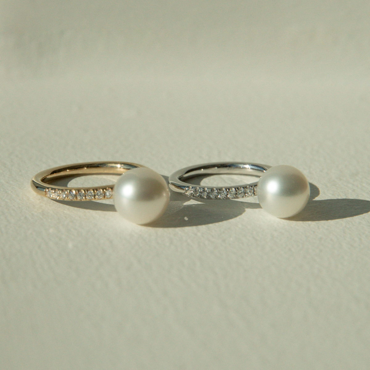 Two pearl rings with diamond accents on a light beige background