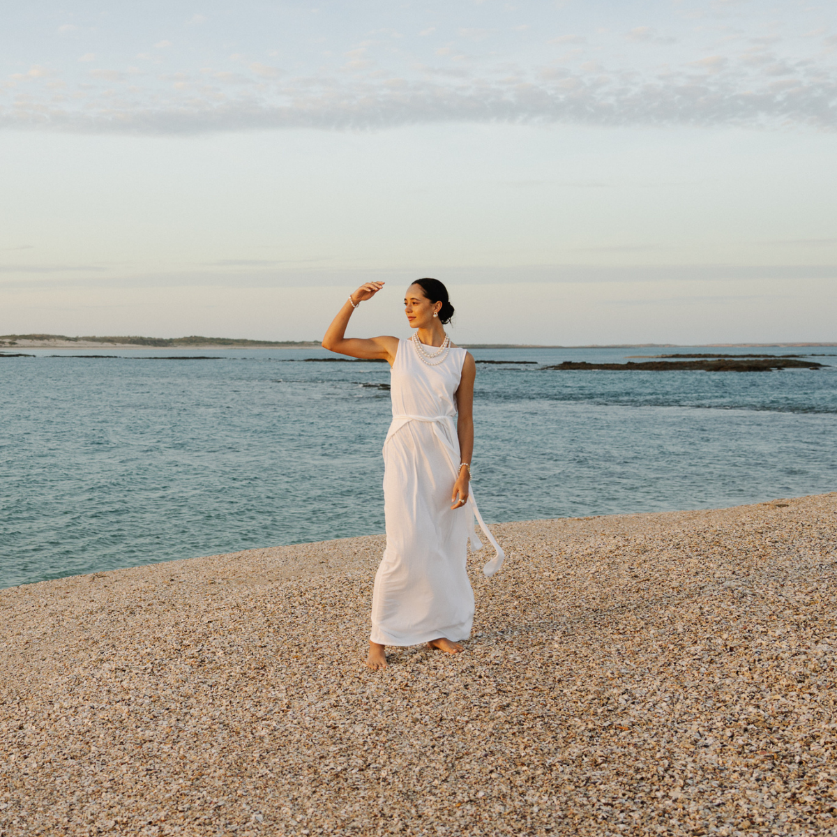 Woman in a white dress standing on a pebbly beach opposite Cygnet Bay Pearl Farm with ocean and sky in the background