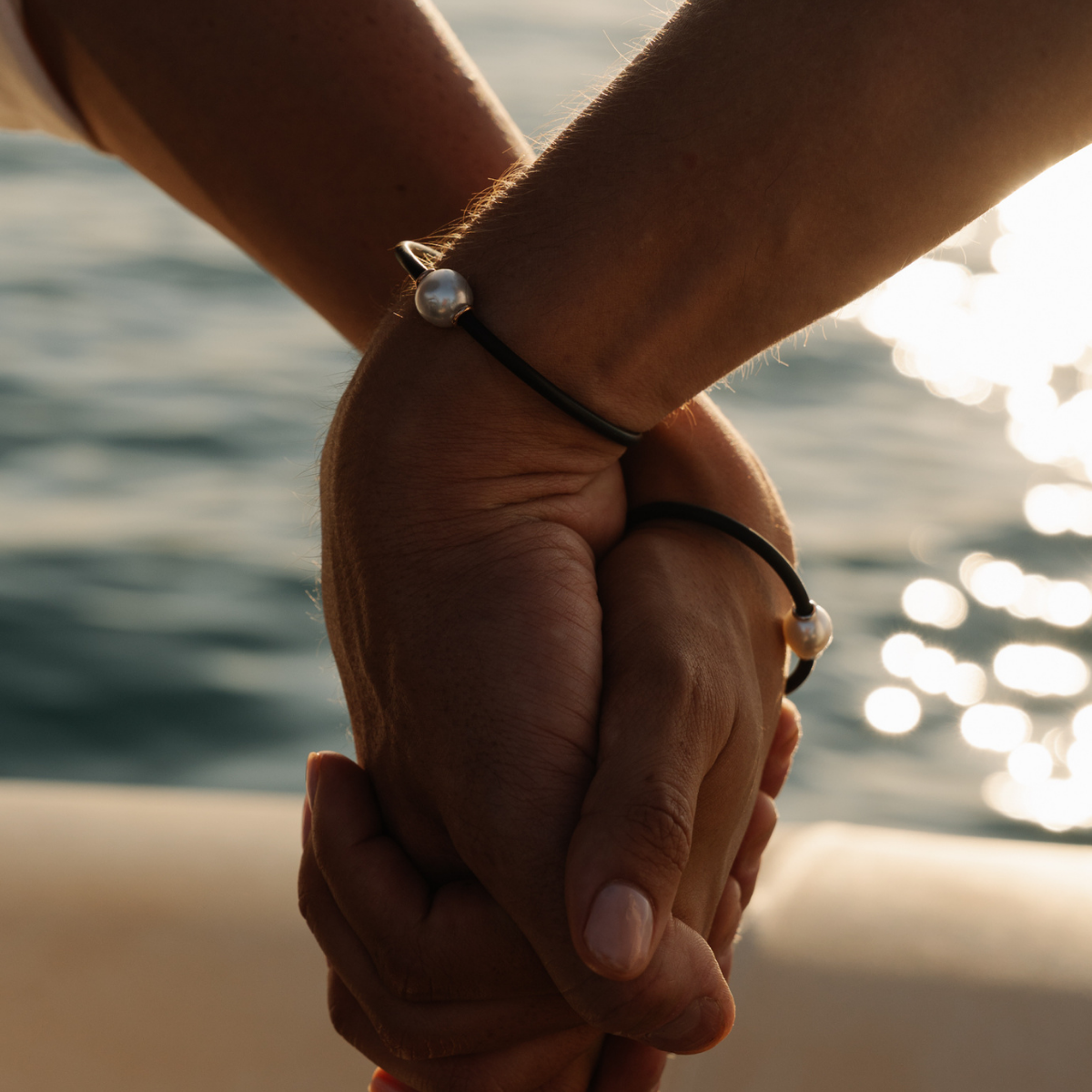 Two hands holding each other, wearing matching neoprene pearl bracelets with a blurred water background