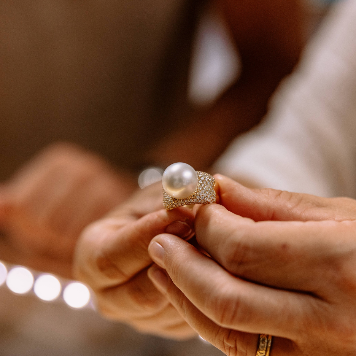 A close-up lifestyle shot of a person holding a ring with a large white pearl and a gold band paved with small white diamonds.