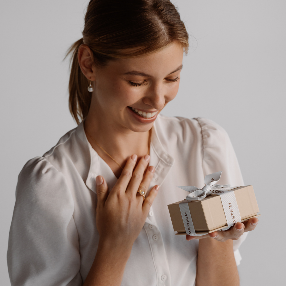 A woman in a white blouse holds a small beige gift box tied with a white ribbon. The box has the brand name 'Pearls of Australia' printed on it.